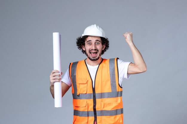 Front close view of young happy construction worker in warning vest with safety helmet and showing blank on gray wall