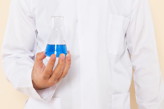 Front close view young male chemist in white special suit holding little flask with blue solution on cream desk science experiment chemistry scientific