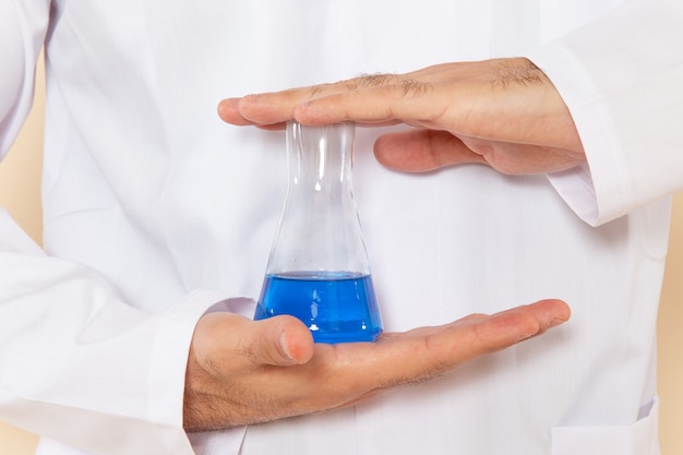 Front close view young male chemist in white special suit holding little flask with blue solution on cream wall science experiment chemistry scientific