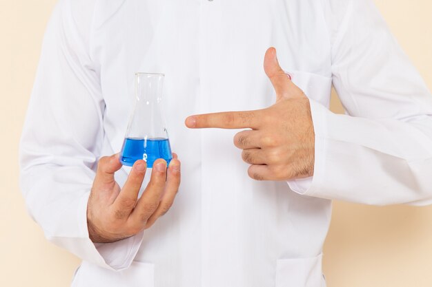 Front close view young male chemist in white special suit holding little flask with blue solution on cream wall science lab experiment chemistry scientific