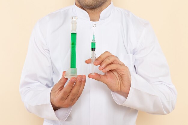Front close view young male chemist in white special suit holding little flask with green solution and injection on cream wall science experiment lab