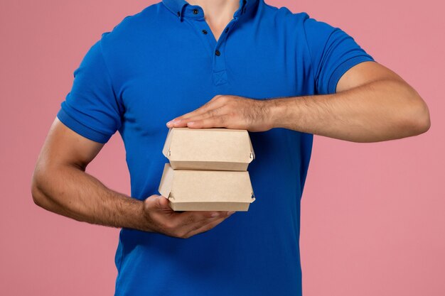 Front close view young male courier in blue uniform cape holding little delivery food packages on the pink wall, service employee delivering