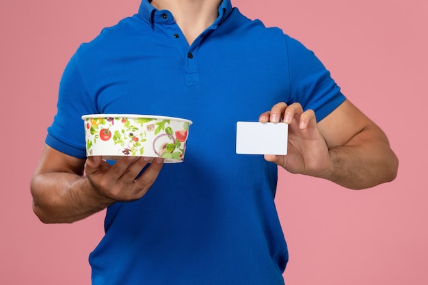 Front close view young male courier in blue uniform cape holding white card and round delivery bowl on light pink wall, uniform delivering service