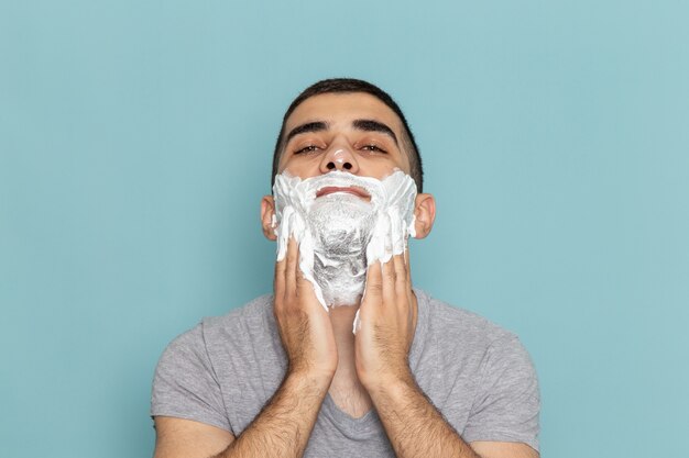 Front close view young male in grey t-shirt covering his face with white foam for shaving on ice-blue wall beard razor shave