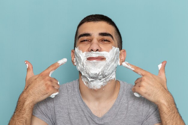 Front close view young male in grey t-shirt covering his face with white foam for shaving on iced-blue desk beard foam razor shave