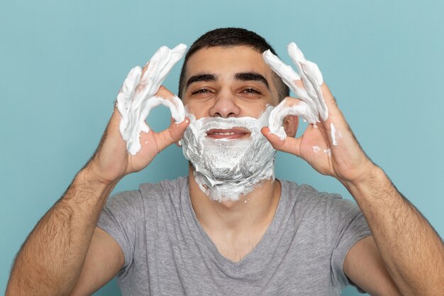 Front close view young male in grey t-shirt covering his face with white foam for shaving showing alright signs on ice-blue wall beard foam shaving male