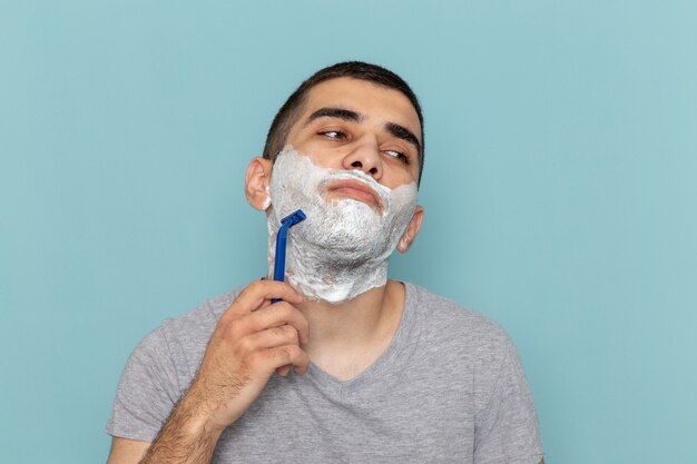 Front close view young male in grey t-shirt shaving his beard with razor on the ice-blue wall beard foam shaving male