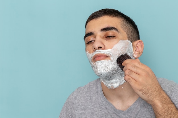 Front close view young male in grey t-shirt with white foam on his face fixing it on ice-blue wall beard foam shaving male