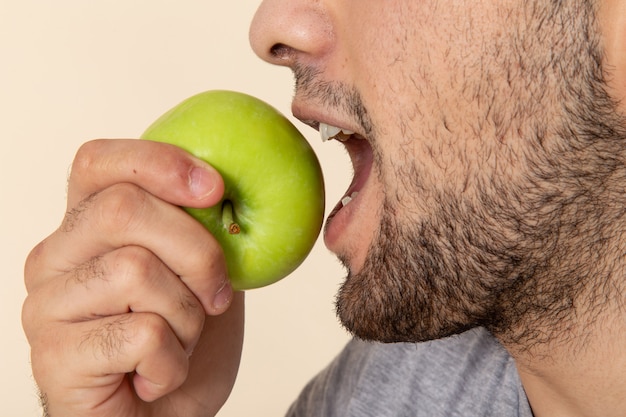 Front close view of young man in grey t-shirt and blue jeans biting green apple