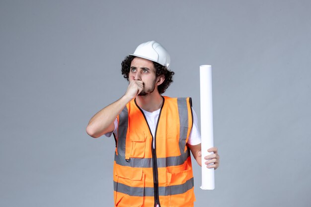 Front close view of young thinking male construction worker in warning vest with safety helmet and showing blank on gray wall