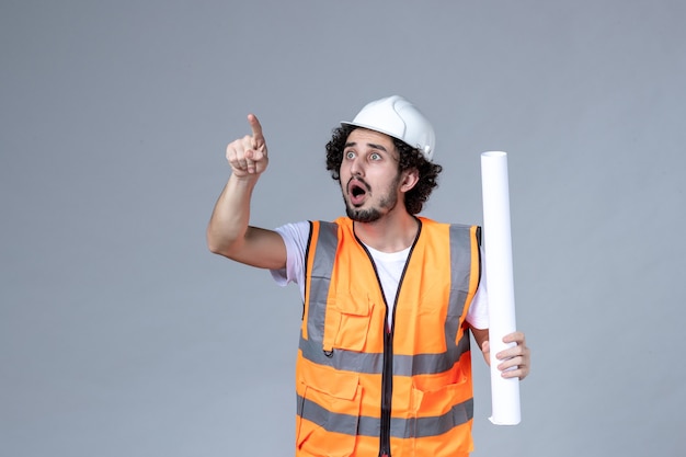 Front close view of young wondering focused male construction worker in warning vest with safety helmet and showing blank on gray wall