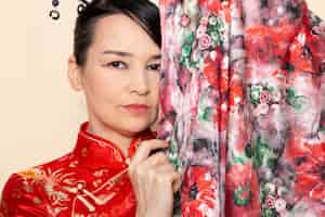 Foto gratuita una geisha giapponese squisita di vista chiusa anteriore in vestito giapponese rosso tradizionale che posa con sorridere elegante del tessuto progettato fiore sulla cerimonia del fondo crema giappone
