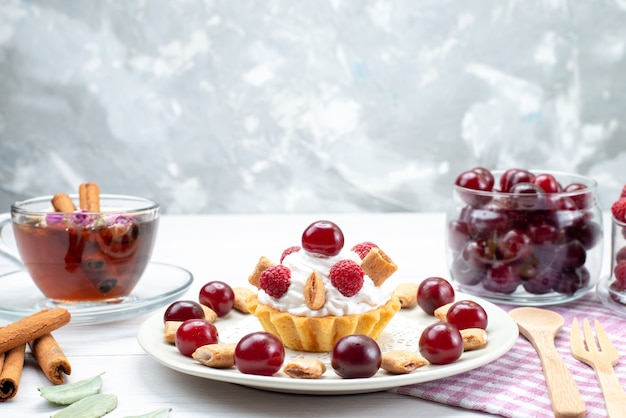 Front closer view little creamy cake with raspberries cherries and little biscuits tea cinnamon on light desk