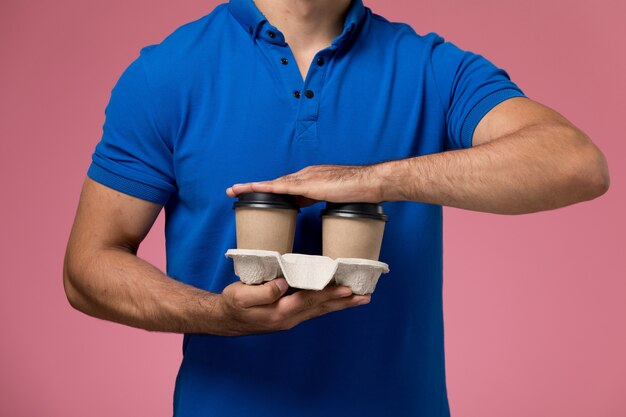 Front closer view male courier in blue uniform holding coffee cups on pink, uniform service delivery worker