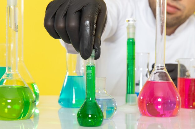 Передний близкий вид молодого мужчины-химика в белом костюме перед столом с цветными растворами, держащего колбу на желтом столе, в научной лаборатории