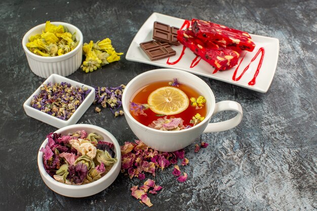 Front shot of a cup of herbal tea with bowls of dry flowers and a plate of chocolate on grey ground