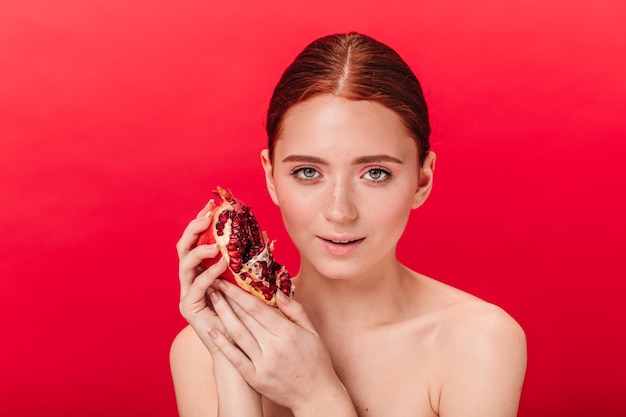 Front view of adorable woman holding ripe garnet. Studio shot of ginger naked girl with pomegranate isolated on red background.