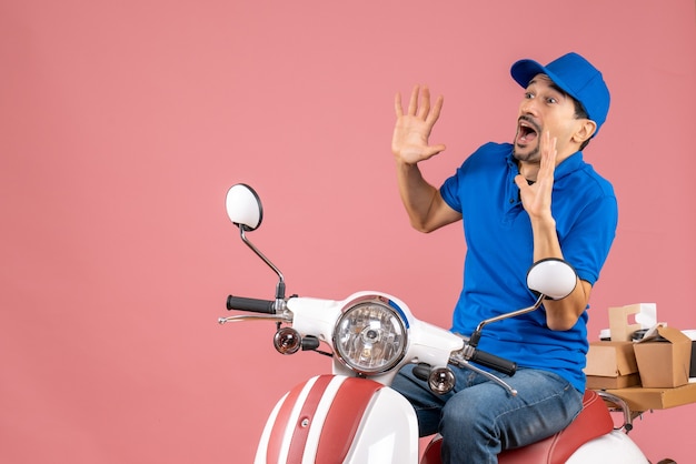 Front view of afraid delivery guy wearing hat sitting on scooter on pastel peach background