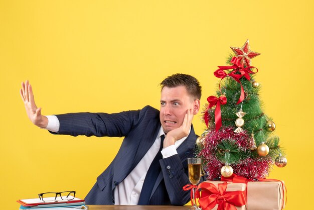 Front view of agitated man trying to stop something sitting at the table near xmas tree and gifts on yellow wall