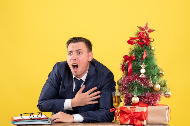 Vista frontale agitato giovane che tiene il petto seduto al tavolo vicino all'albero di natale e regali su sfondo giallo