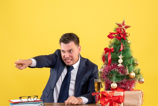 Front view of aguitated young man pointing at something sitting at the table near xmas tree and gifts on yellow wall