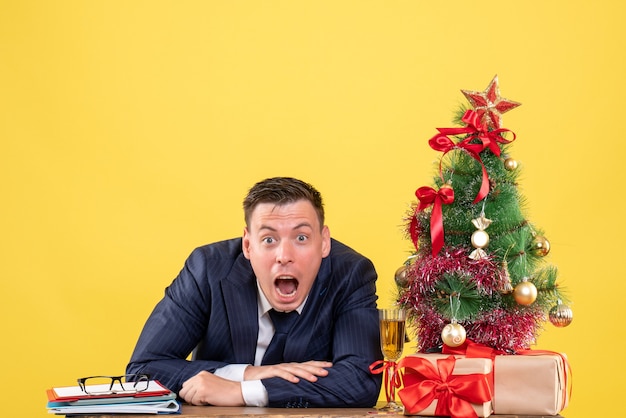 Front view amazed man opening his mouth sitting at the table near xmas tree and presents on yellow background