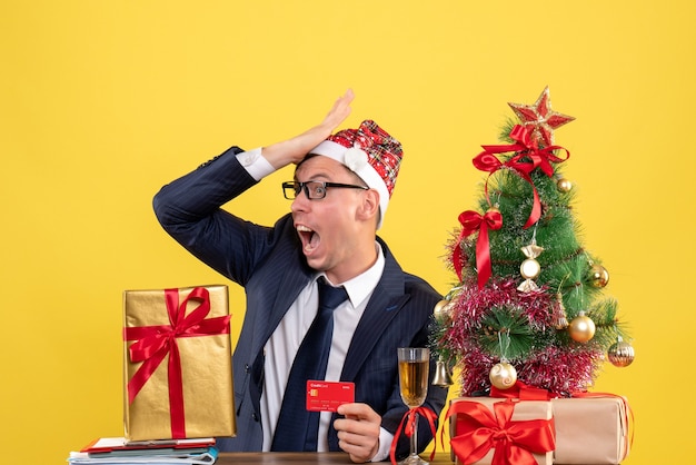 Front view of amazed man putting hand to his forehead sitting at the table near xmas tree and presents on yellow wall