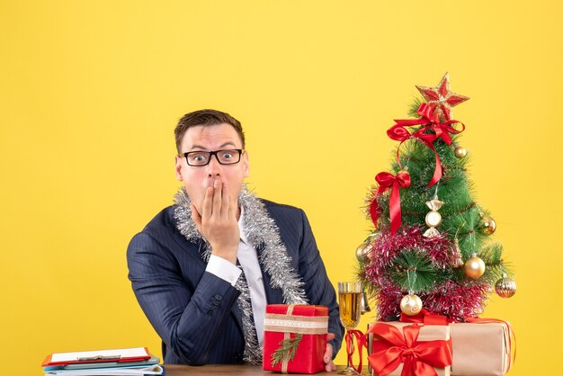 Вид спереди изумленный мужчина, прикладывающий руку ко рту, сидя за столом возле рождественской елки и подарки на желтом фоне