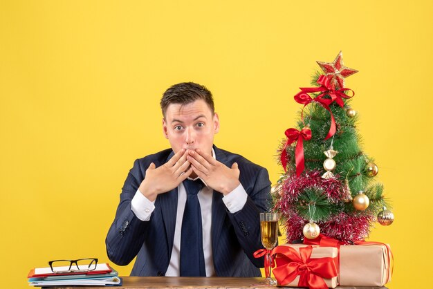 크리스마스 트리 근처 테이블에 앉아 그의 입에 손을 넣어 놀란 남자의 전면보기 노란색 벽에 선물
