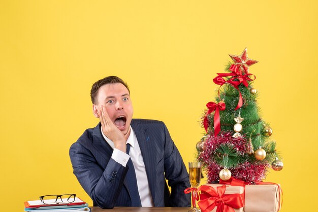 Front view of amazed man shouting while sitting at the table near xmas tree and presents on yellow wall