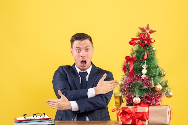 크리스마스 트리 근처 테이블에 앉아 방향을 보여주는 놀란 남자의 전면보기와 노란색 벽에 선물
