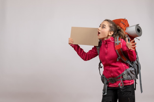 Front view of amazed pretty woman with big backpack holding up cardboard