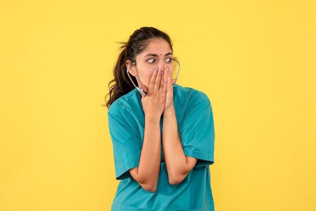 Vista frontale stupita donna medico in uniforme che mette entrambe le mani sul viso su sfondo giallo