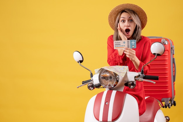 Front view of amazed young lady in red dress holding up ticket on moped