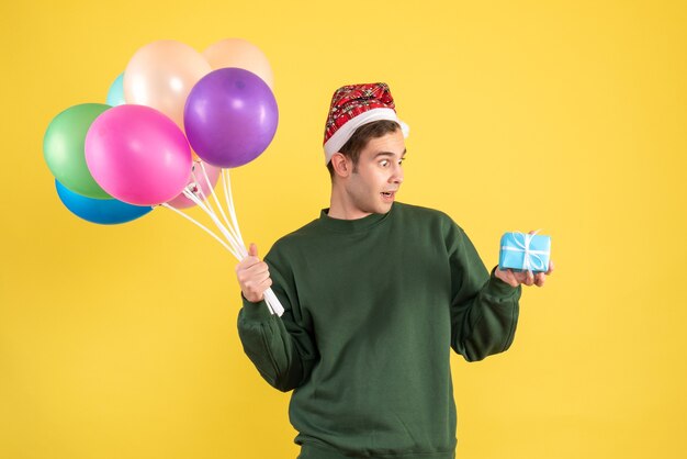 Front view amazed young man with santa hat and colorful balloons standing on yellow 