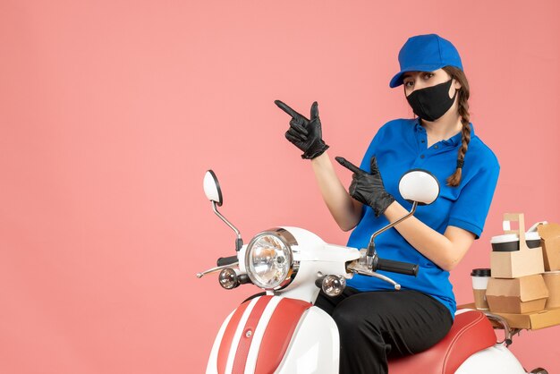 Free photo front view of ambitious courier girl wearing medical mask and gloves sitting on scooter delivering orders on pastel peach background