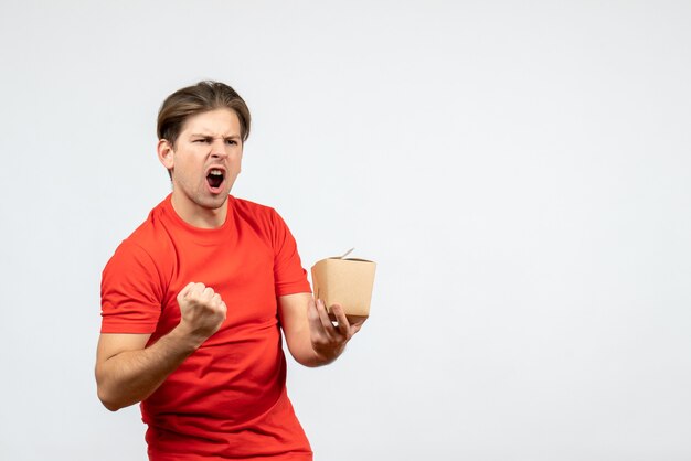 Front view of ambitious and emotional young guy in red blouse holding small box on white background