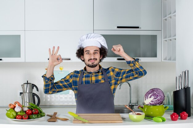 新鮮な野菜とキッチンツールで調理し、白いキッチンで彼の筋肉を示す眼鏡ジェスチャーを作る野心的な男性シェフの正面図