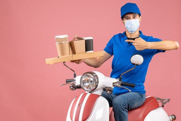 Front view of ambitious male delivery person in mask wearing hat sitting on scooter delivering orders on pastel peach background