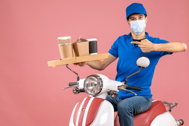 Вид спереди амбициозного доставщика-мужчины в маске в шляпе, сидящего на скутере, доставляющего заказы на пастельно-персиковом фоне