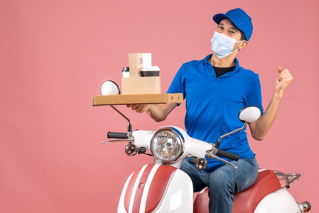 Вид спереди амбициозного доставщика-мужчины в маске в шляпе, сидящего на скутере, доставляющего заказы на пастельно-персиковом фоне