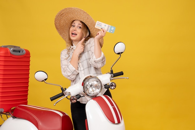 Front view of ambitious young woman wearing hat collecting her luggage sitting on motorcycle and showing ticket