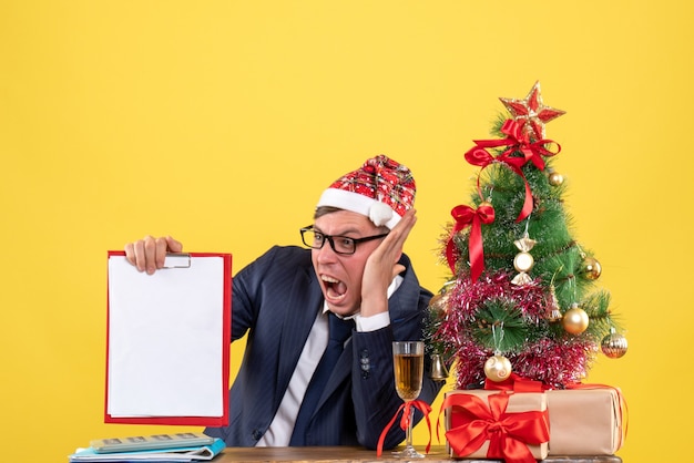 Front view angry business man sitting at the table near xmas tree and presents on yellow background