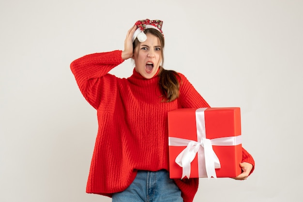 Front view angry girl with santa hat holding present holding her head standing