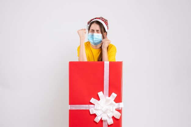 Front view angry girl with santa hat showing punches standing behind big xmas gift