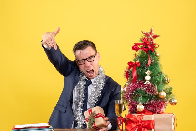 Вид спереди сердитый человек, указывая пальцем вниз, сидит за столом возле рождественской елки и представляет на желтом фоне