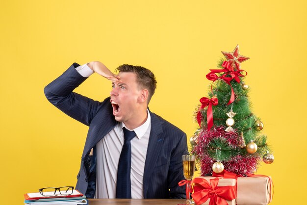 Front view of angry man observing sitting at the table near xmas tree and presents on yellow wall