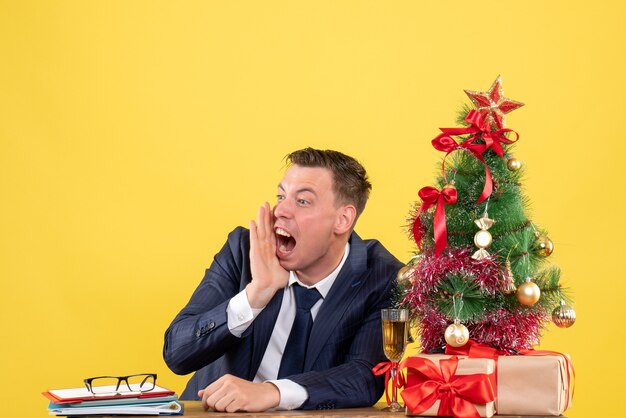 Вид спереди сердитый человек кричит, сидя за столом возле рождественской елки и представляет на желтом фоне