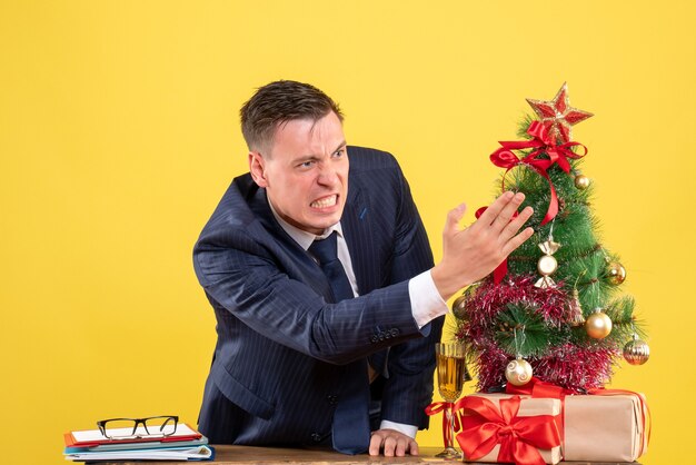 Front view of angry man standing behind the desk near xmas tree and presents on yellow wall