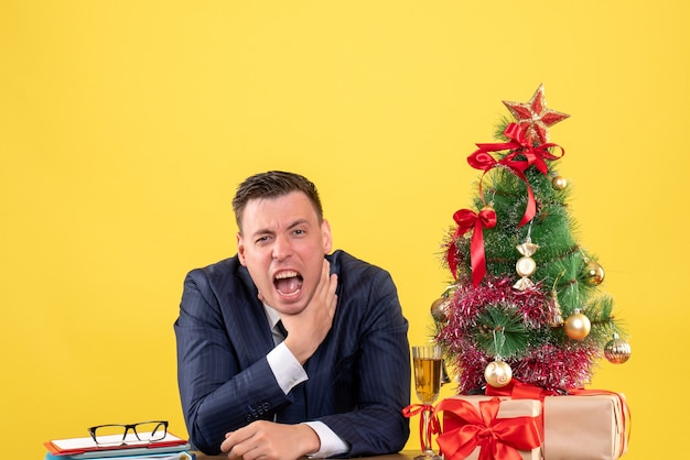 Front view angry man strangling himself with hand sitting at the table near xmas tree and gifts on yellow background